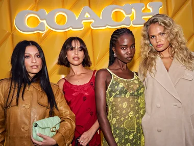 Four women posing in front of a yellow background with the illuminated Coach logo, wearing fashionable outfits.